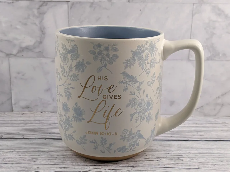 A religious mug that's a cream color with a light blue flower design and a gold Bible verse with a blue interior.