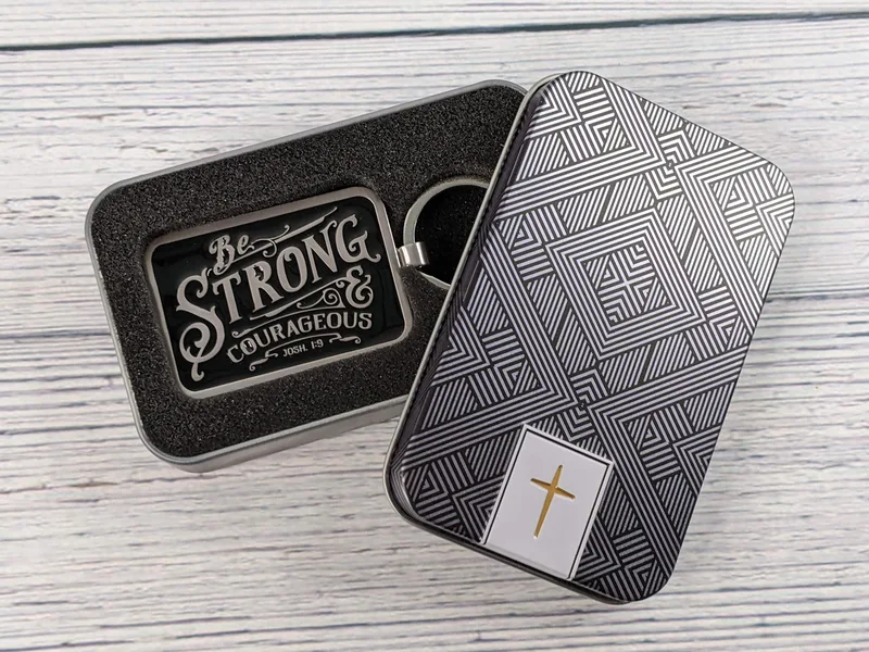 A silver metal Bible verse keyring with a black enamel design and the verse Joshua 1:9, that comes in a gift tin.