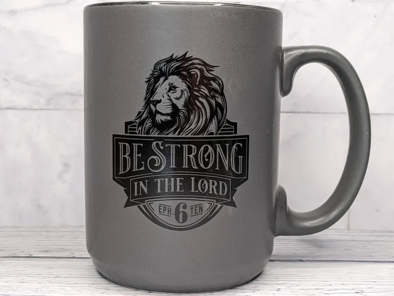 A matte gray religious coffee mug with an image of a lion and a Bible verse: Be strong in the Lord.