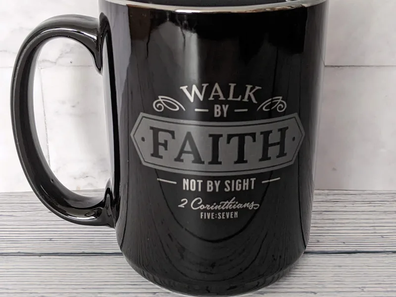 A glossy black mug with a Bible verse: Walk by faith not by sight.