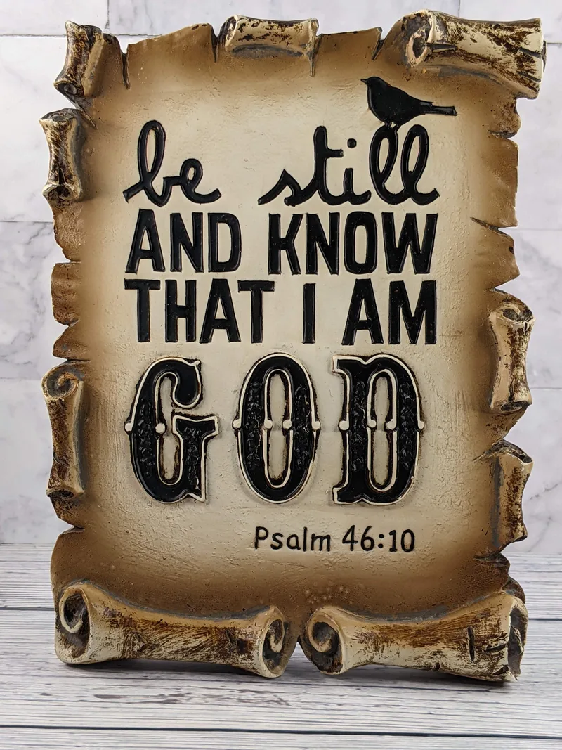 A tan and brown resin Christian shelf-sign, shaped like an opened scroll with a Bible verse from Psalm 46:10, "Be still and know that I am God."