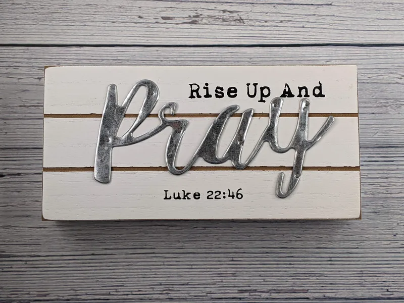A white home decor wall sign with metal lettering and a "Rise up and pray -Luke 22:46" inscription.