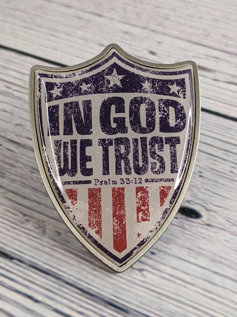 A chrome metal visor clip shaped like a shield with stars, stripes, and the words, "In God we trust."