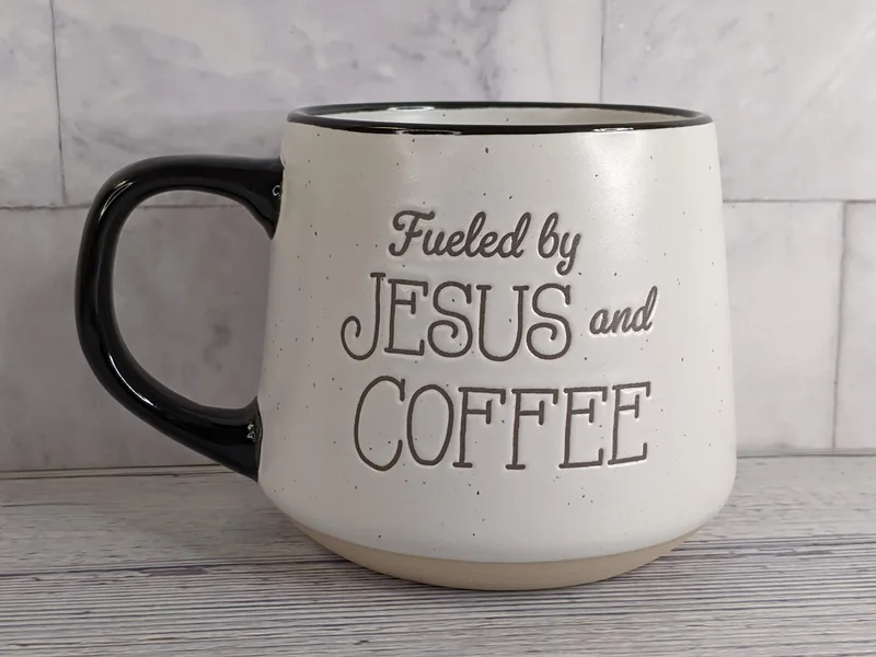 An off-white stoneware coffee mug with a black handle and the words "Fueled by Jesus and Coffee" on both sides.