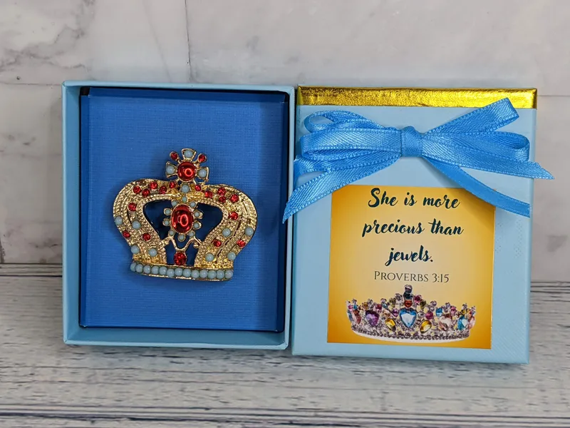 A gold crown pin with red and light blue faux jewels in a blue gift box with a Proverbs 3:15 Bible verse on the lid.