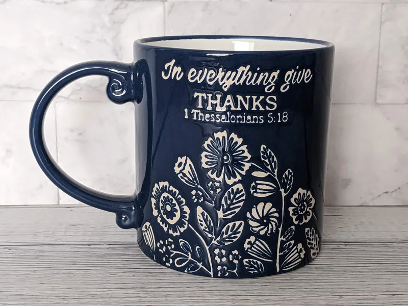 A blue Christian Scripture mug with a tan floral design and the words "In everything give thanks. 1 Thessalonians 5:18."