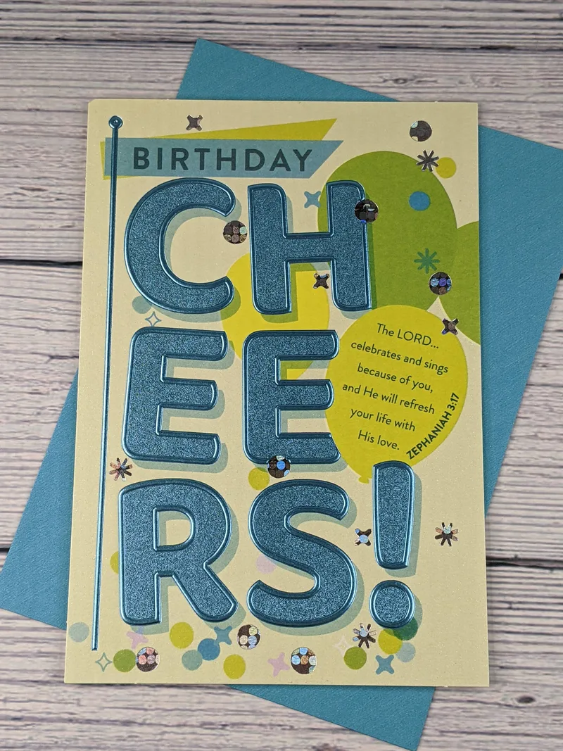 A pale green greeting card with metallic blue words "Birthday Cheers!" and a Bible verse on an image of a balloon.