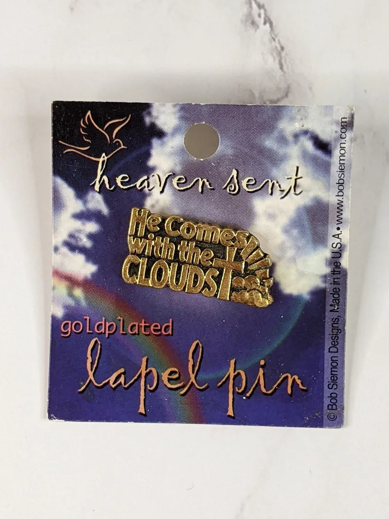 A gold metal lapel pin with a cross design and the words, "He comes with the clouds."