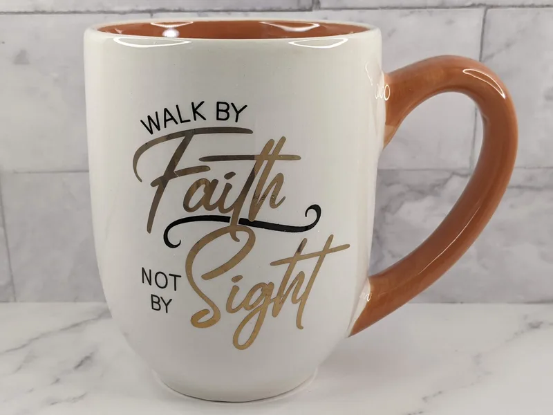 A white coffee mug with a coral-colored handle and the Bible verse message to walk by faith.