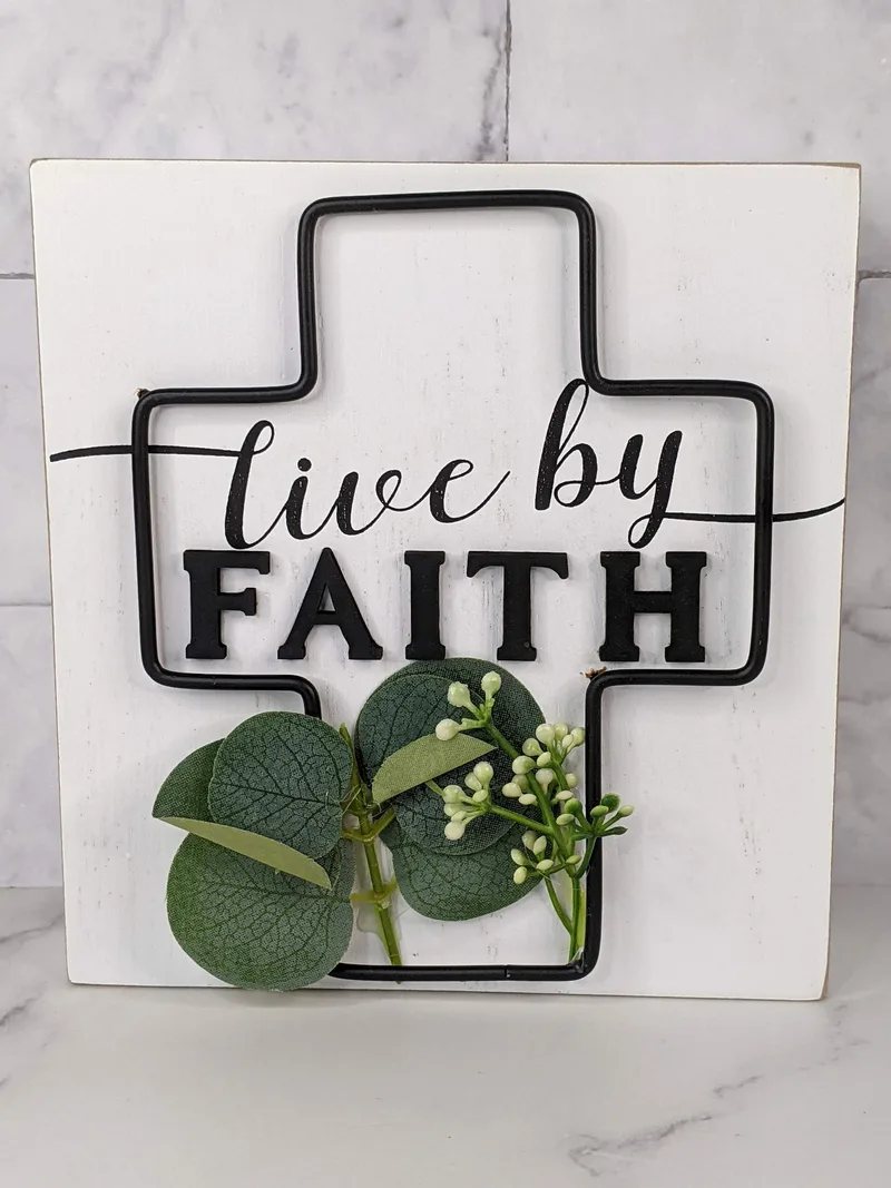 A religious white sign with a cross symbol, embellished with leaves, and the words "live by faith."